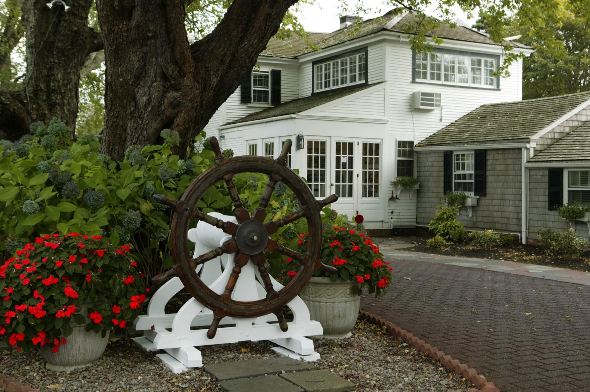 Exterior view of Captain's House Inn with brick drive, tall mature trees and a captain's wheel flanked by red flowers